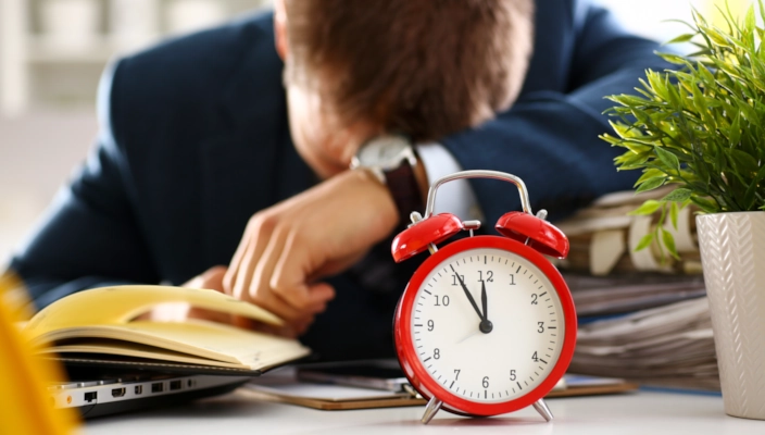 Person liegt hinter analoger Uhr mit Kopf auf dem Tisch, steht unter Zeitdruck
